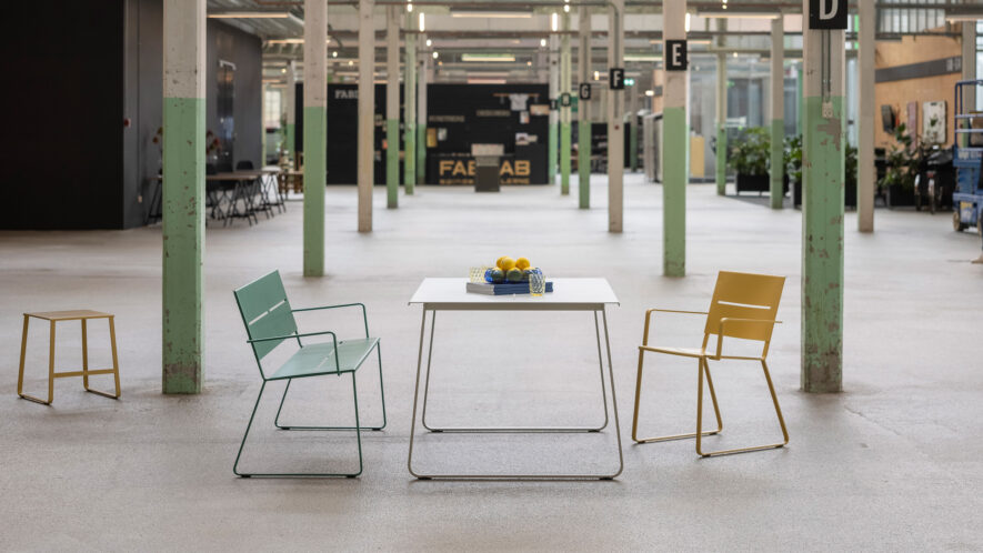 White metal table, yellow metal chair, green metal park bench and sand coloured metal stool is seen in an industrial interior